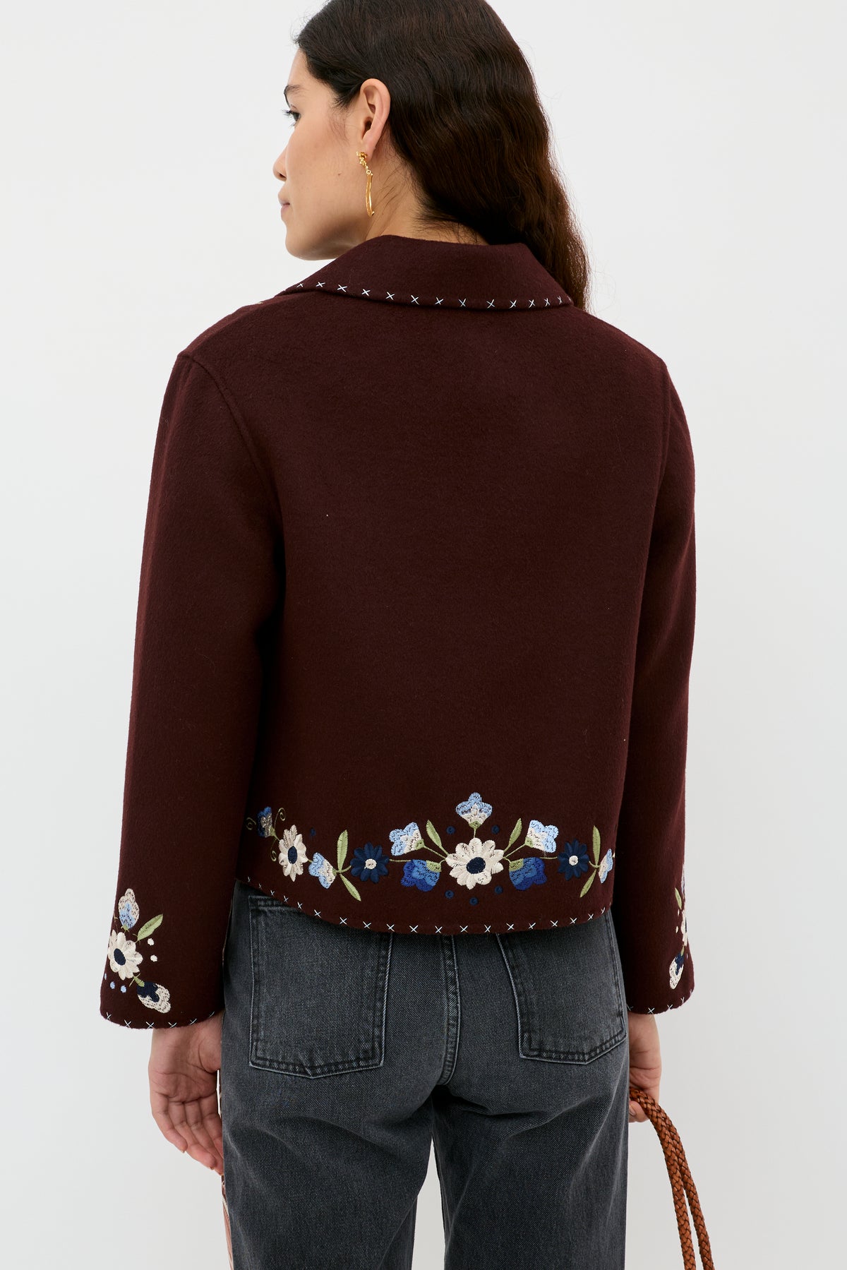 A woman with wavy dark hair and gold hoop earrings stands facing away, wearing the Jude - Bliss Embroidery Brown jacket by RIXO with detailed embroidery at the hem and sleeves, styled with a brown woven bag and dark jeans.