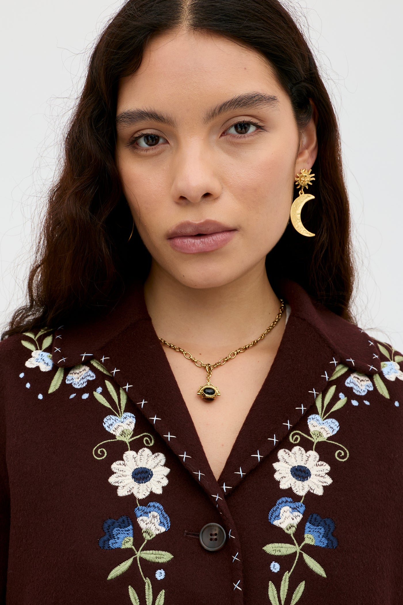 A woman with long brown hair wears the Jude - Bliss Embroidery Brown top featuring blue and white floral RIXO embroidery, paired with a gold eye pendant necklace and a gold crescent moon earring. She faces forward with a neutral expression.