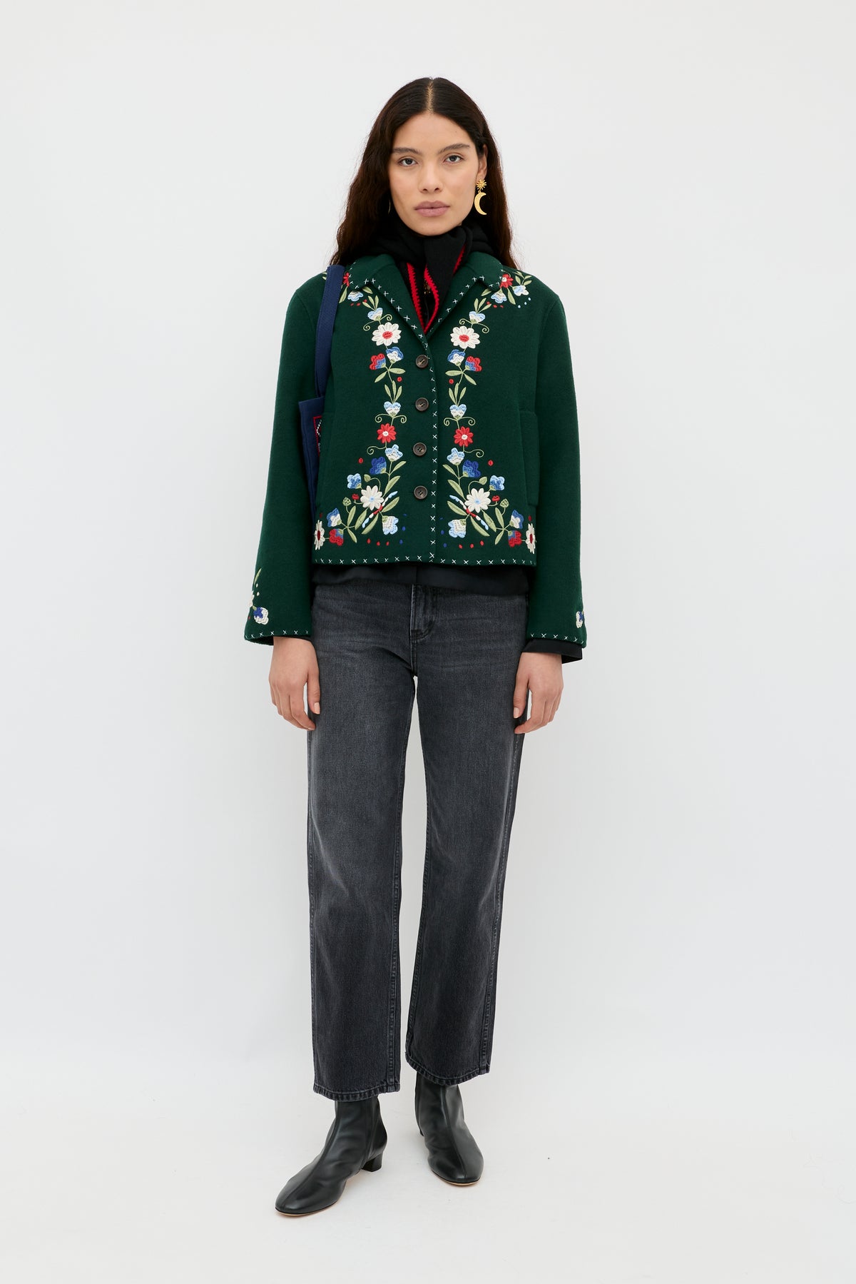 A woman stands against a white background wearing the Jude - Bliss Embroidery Green jacket, styled with black jeans, black ankle boots, and gold earrings. She has long dark hair and a neutral expression.