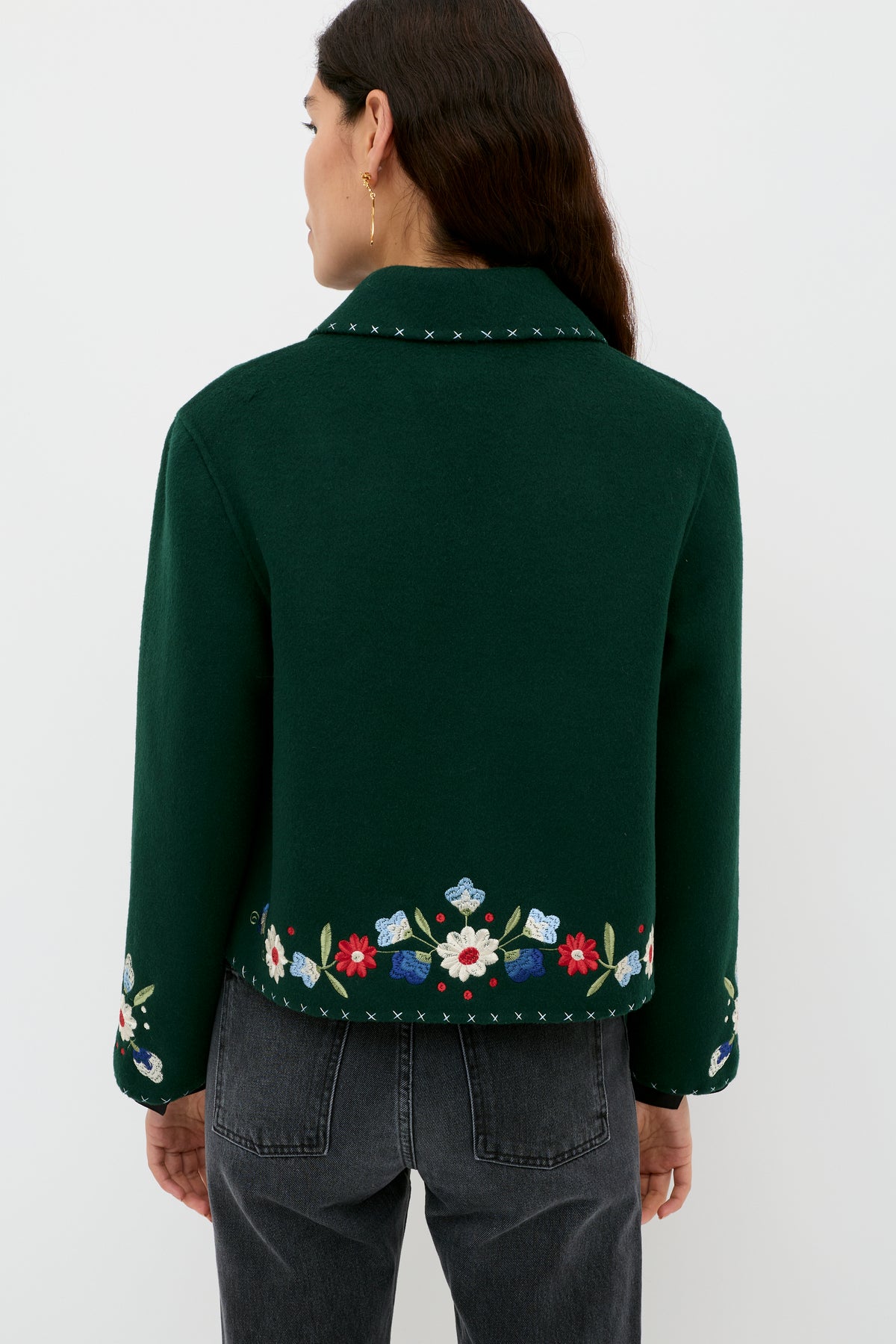 A woman with long brown hair wears the Jude - Bliss Embroidery Green jacket, showcasing its colorful embroidered hem and cuffs. She pairs it with black jeans and faces away, highlighting the back of the jacket against a plain white background.
