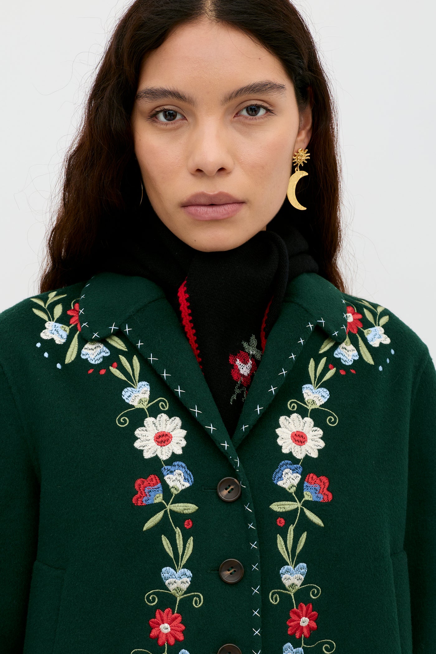 A woman with long brown hair wears the dark green Jude - Bliss Embroidery Green jacket, featuring colorful embroidery, a black scarf, and crescent moon-shaped earrings, looking directly at the camera against a plain background.