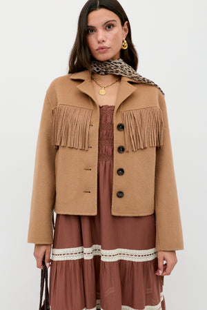 A woman wears the Ashleigh - Camel jacket with fringe over a mauve lace-accented dress, accessorized with a leopard print scarf and layered gold necklaces, standing against a plain white background.