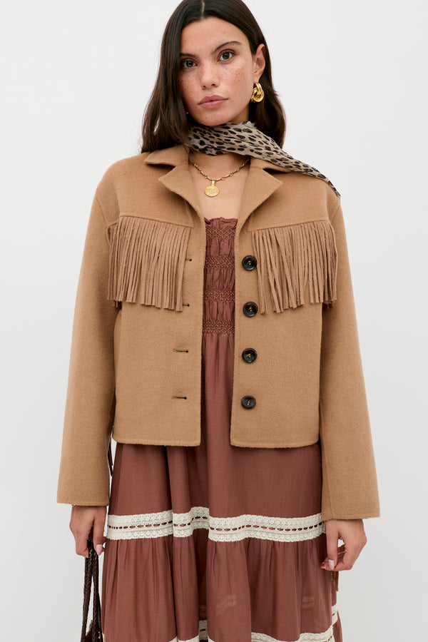 A woman wears the Ashleigh - Camel jacket with fringe over a mauve lace-accented dress, accessorized with a leopard print scarf and layered gold necklaces, standing against a plain white background.