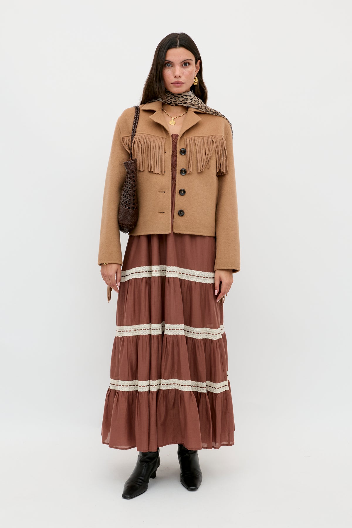 A woman stands against a plain background wearing the Ashleigh - Camel jacket with fringe, a brown tiered maxi dress with lace trim, a printed scarf, black ankle boots, and carrying a brown textured shoulder bag.