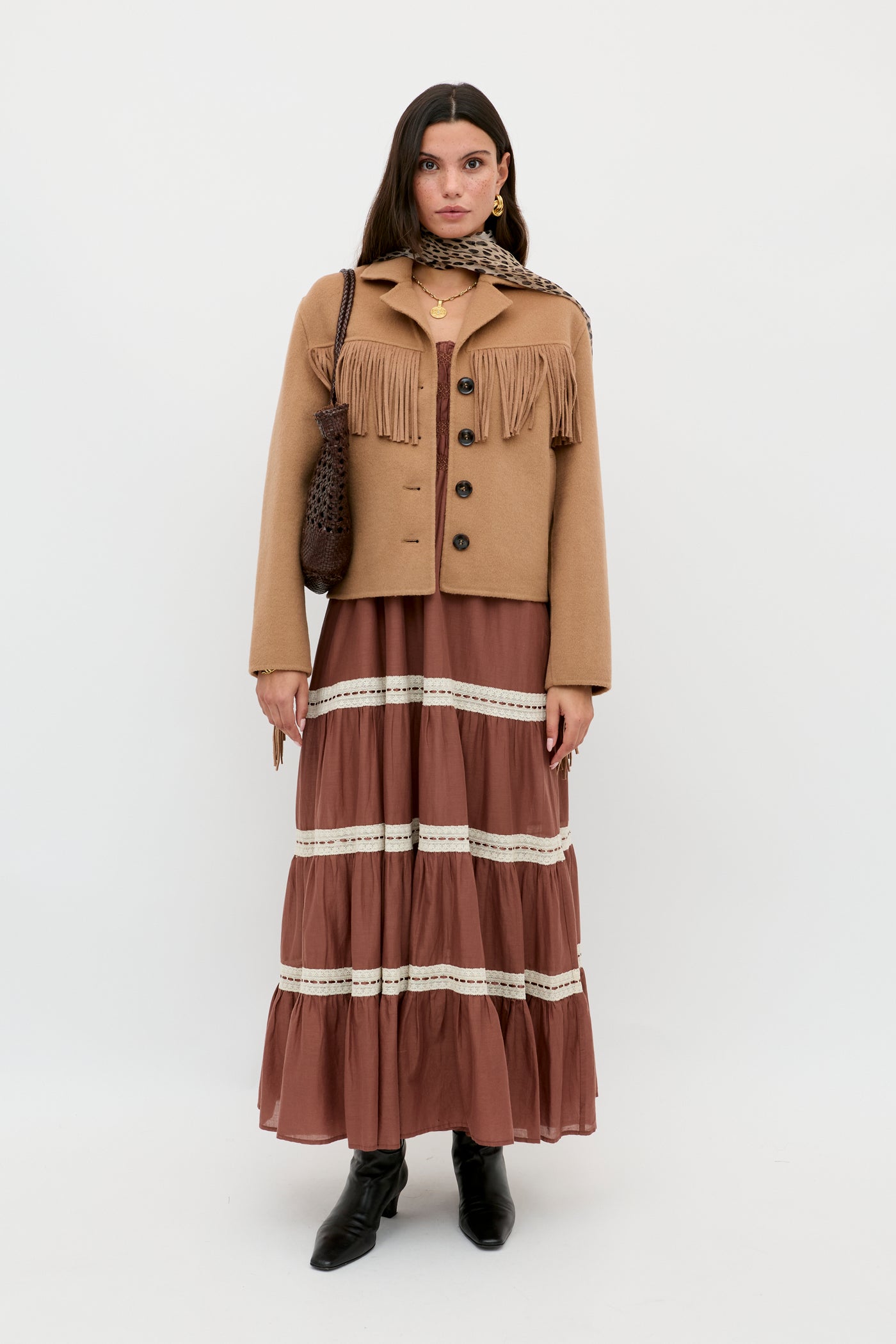 A woman stands against a plain background wearing the Ashleigh - Camel jacket with fringe, a brown tiered maxi dress with lace trim, a printed scarf, black ankle boots, and carrying a brown textured shoulder bag.