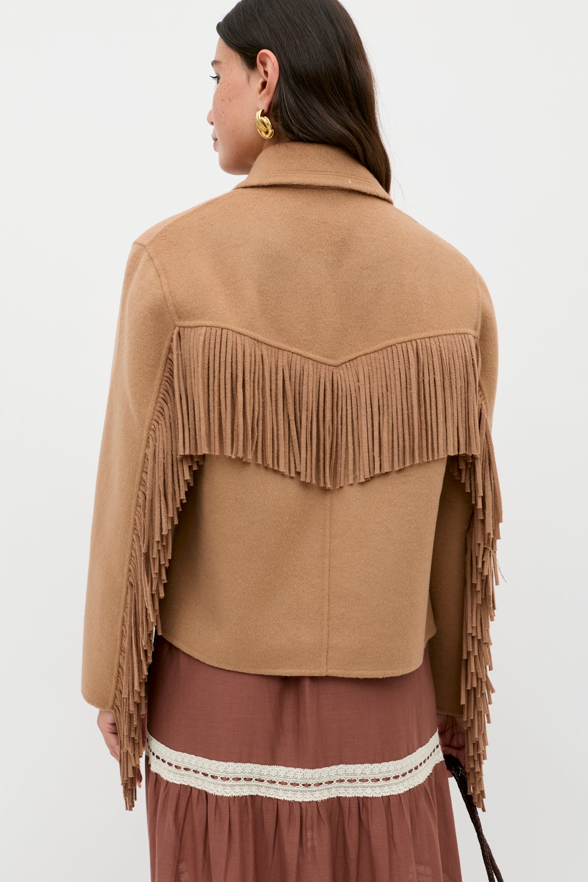 A woman with long brown hair faces away, wearing the Ashleigh - Camel jacket with fringe across the back and sleeves, paired with a mauve skirt trimmed in white lace and accessorized with a gold hoop earring.