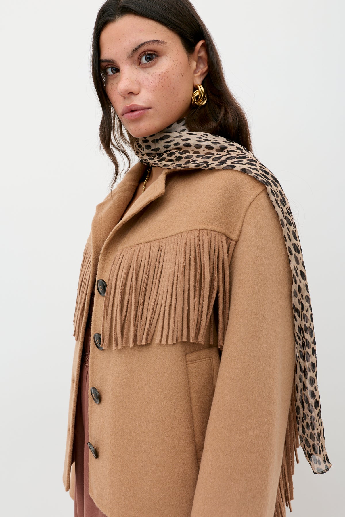 A woman with long brown hair and gold earrings models the Ashleigh - Camel jacket with a leopard print scarf over her shoulder, standing against a plain white background.
