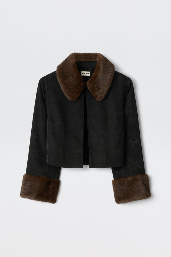 The Luanne - Black is a cropped jacket with a subtle floral pattern, wide brown faux fur collar, and matching cuffs, shown against a plain white background.