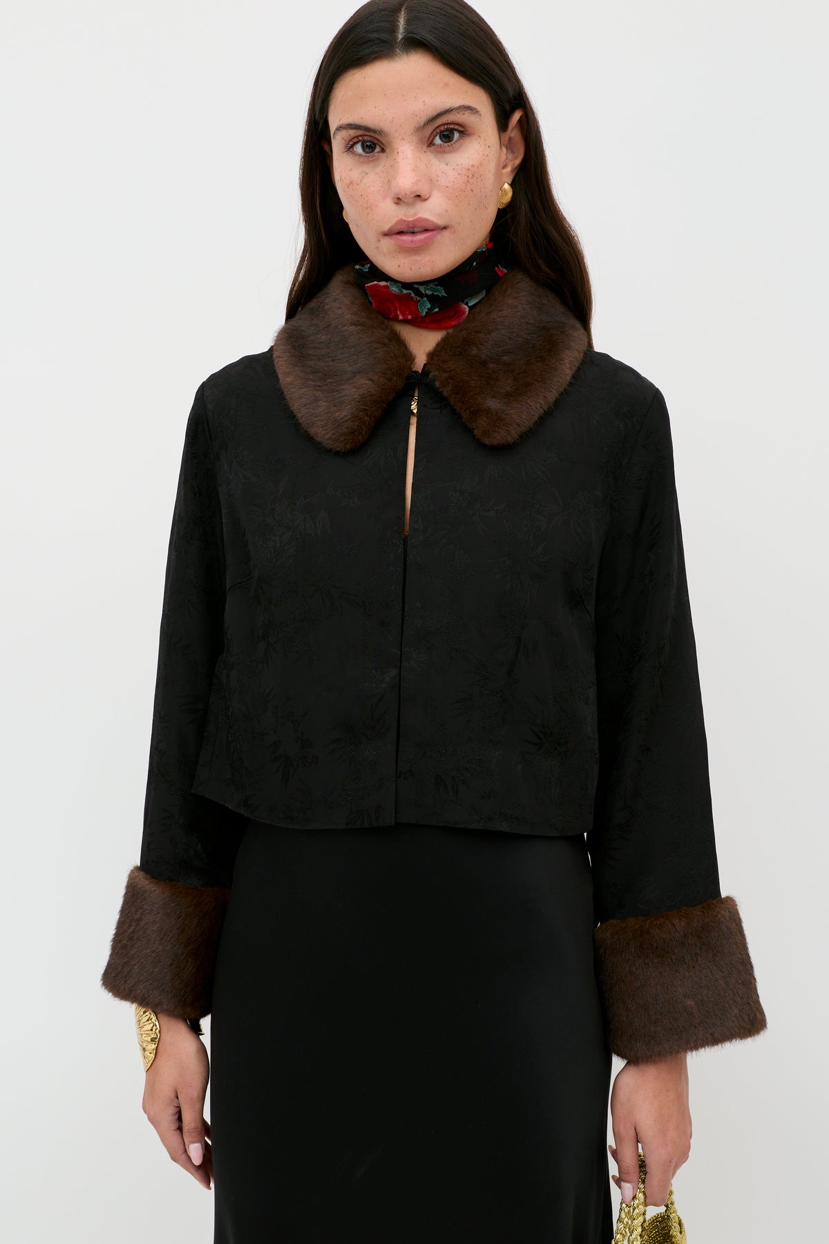 A woman stands against a plain background, wearing the Luanne - Black jacket with a brown fur collar and fur-trimmed cuffs. She pairs it with gold earrings, a floral neck scarf, long dark hair, and holds a small yellow clutch.