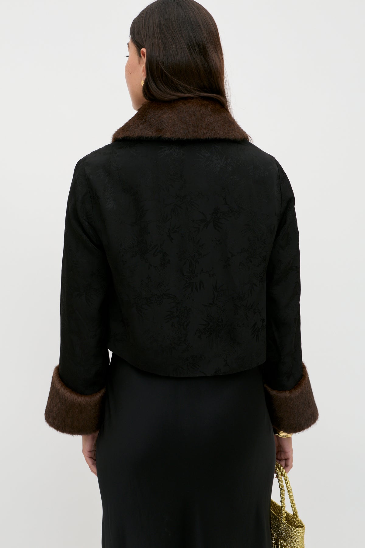 A woman with long brown hair, seen from the back, wears the Luanne - Black jacket with a subtle floral pattern, brown faux fur trim on the collar and cuffs, paired with a black satin skirt and holding a gold purse in her left hand.