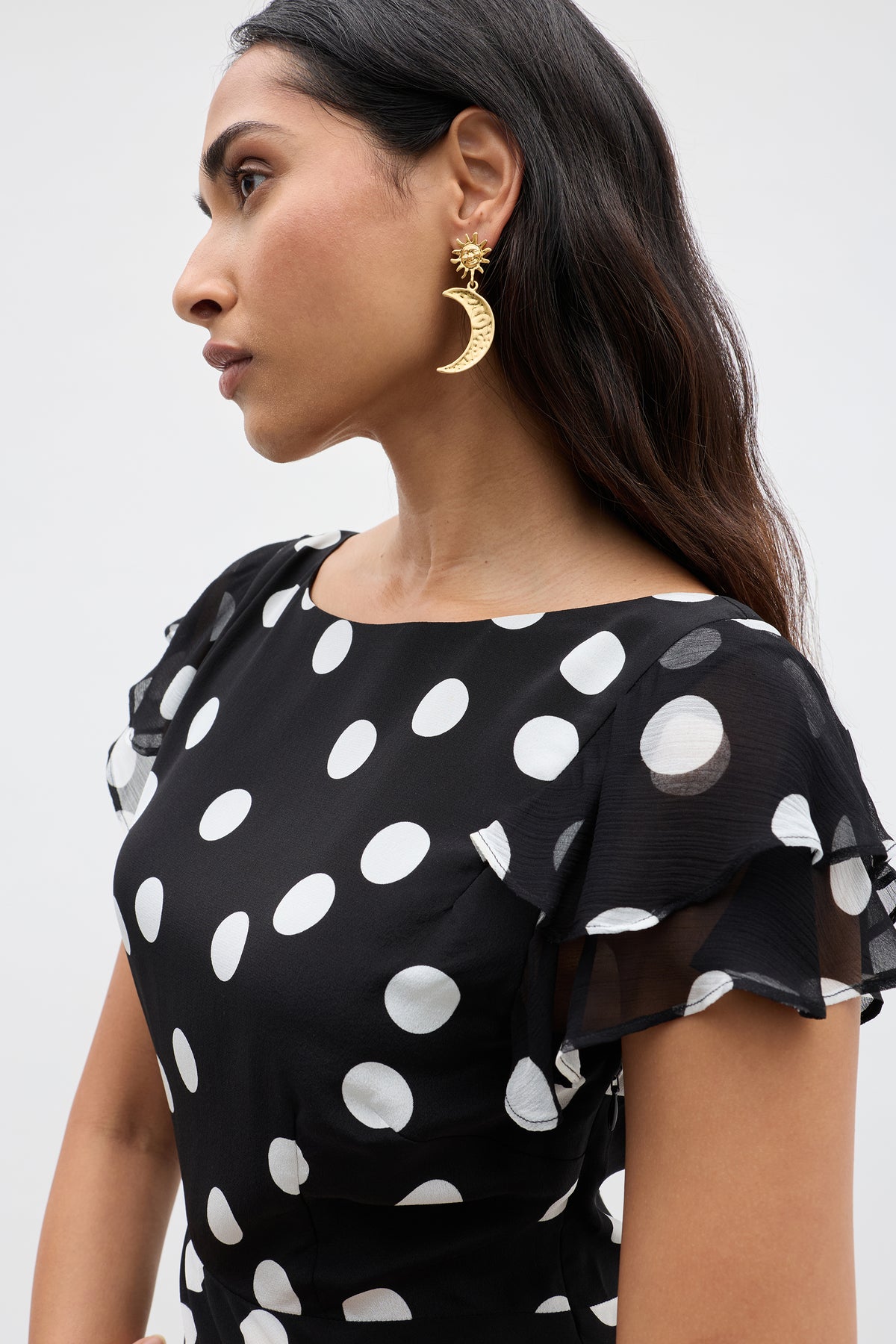 A woman in a black dress with large white polka dots and ruffled sleeves, wearing the Davies - Gold vintage-style celestial earrings, poses against a plain light background.