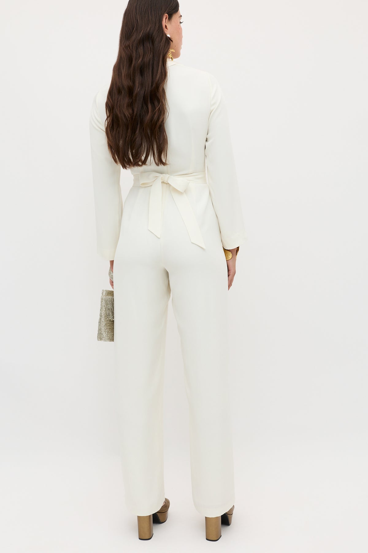 A woman with long brown hair, seen from behind, wears the emilia - ivory seventies-inspired jumpsuit with long sleeves and a waist tie. She holds a small textured clutch and stands on light brown platform heels against a plain background.
