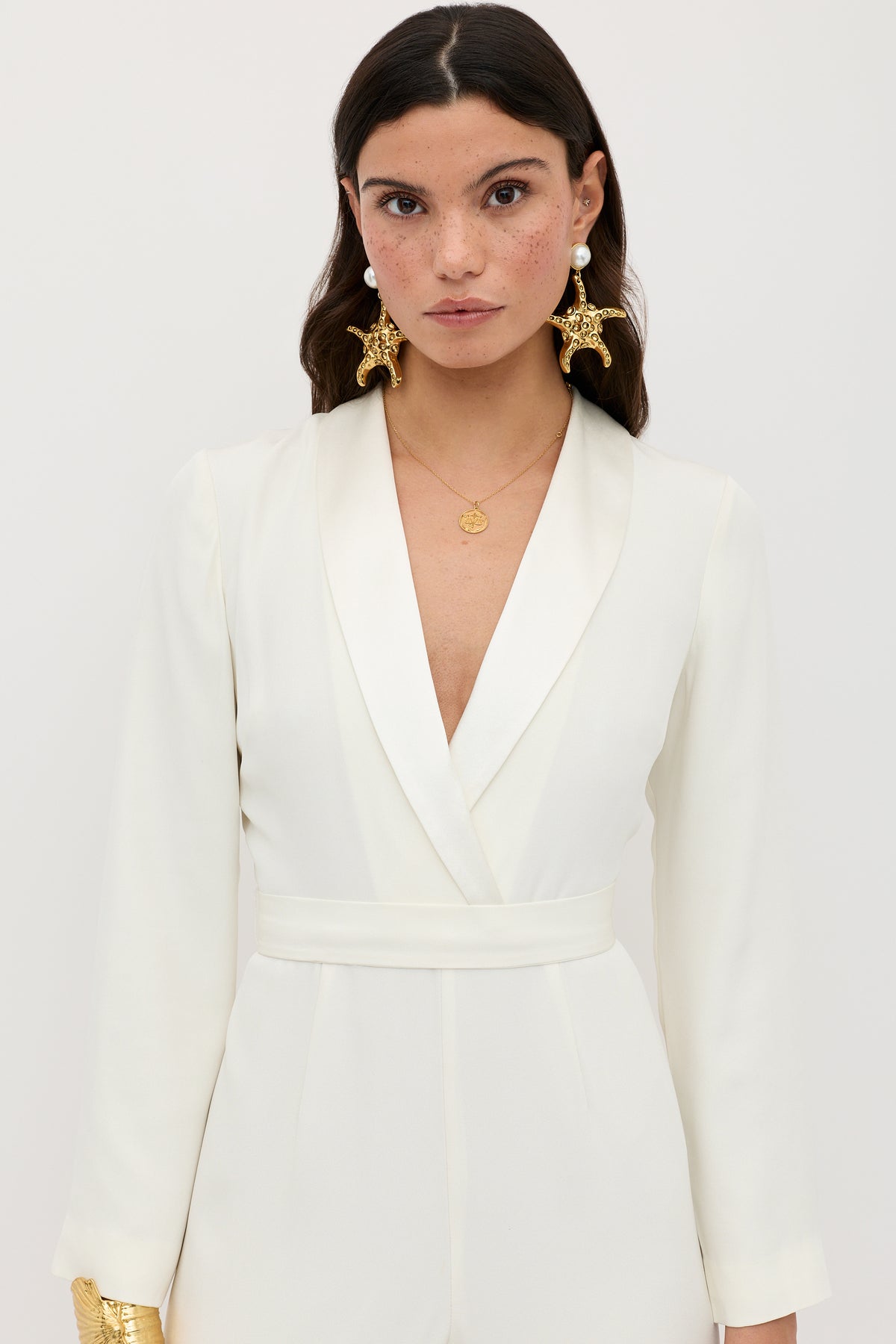 A woman with long brown hair models the emilia - ivory, a tailored white blazer dress with a plunging V-neckline, paired with gold sea-star earrings and a gold pendant necklace, against a plain white background.