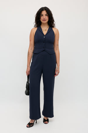 A woman with wavy dark hair and visible arm tattoos stands against a white background, dressed in the Sienna 2 – Navy sleeveless vest and matching pants. She accessorizes with a black handbag and black heels.