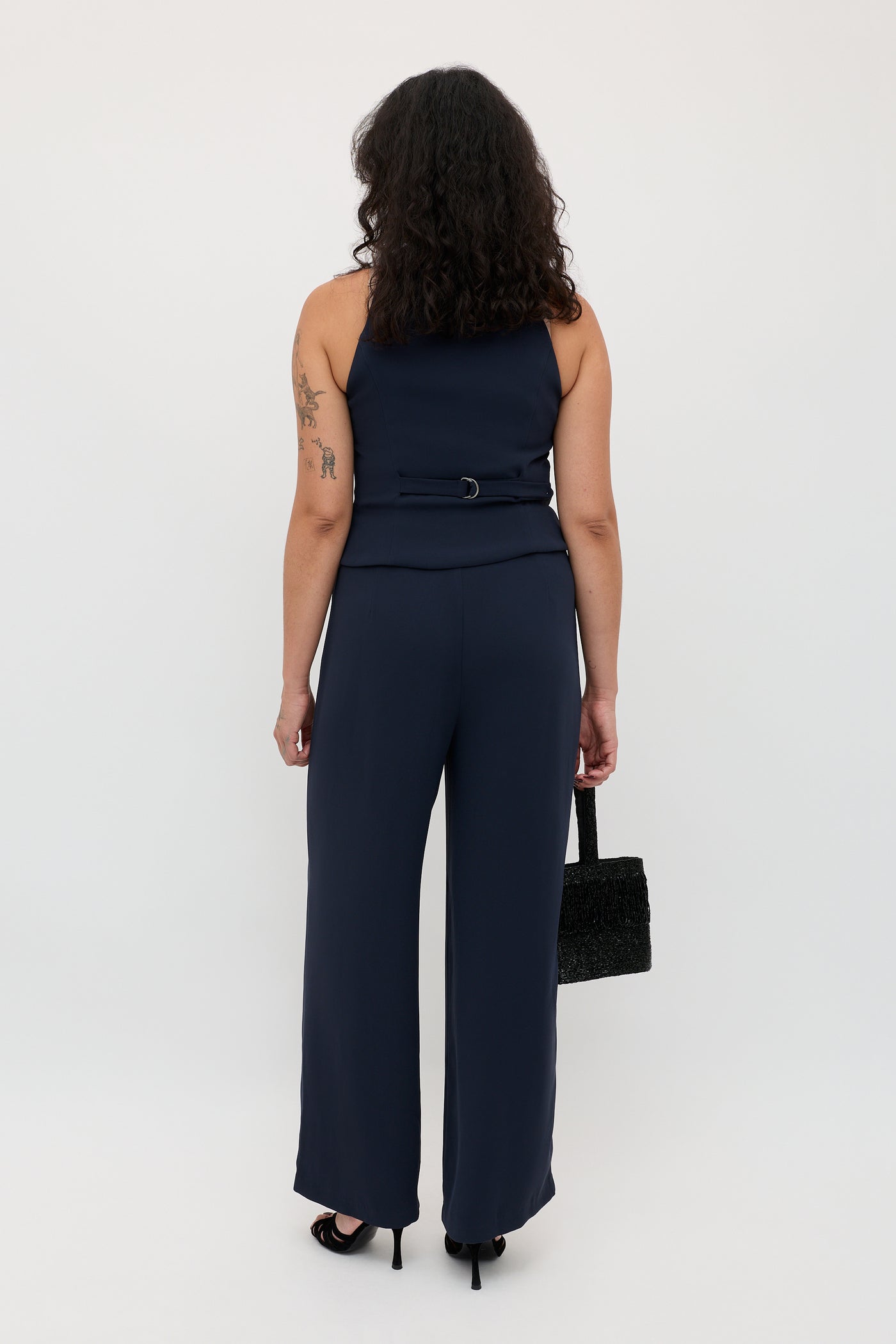 A woman with curly hair and a tattoo on her left arm stands facing away, wearing the Sienna 2 - Navy sleeveless jumpsuit paired with black heels and carrying a black handbag.
