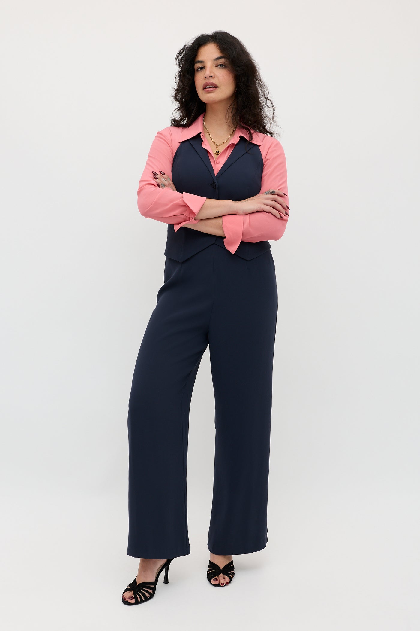 A woman stands with arms crossed against a plain white background, wearing the Sienna 2 - Navy vest and matching navy pants, paired with a pink shirt and black heels.