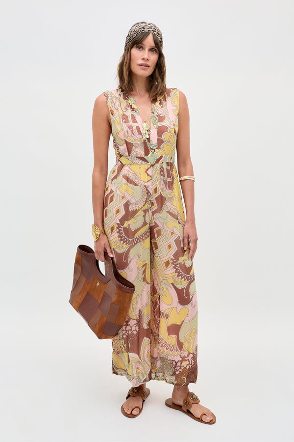 Wearing the Brigid - Bianca Abstract Green sleeveless maxi dress, tan sandals, a beaded headband, gold jewelry, and holding a brown leather bag, a woman stands against a plain light background.