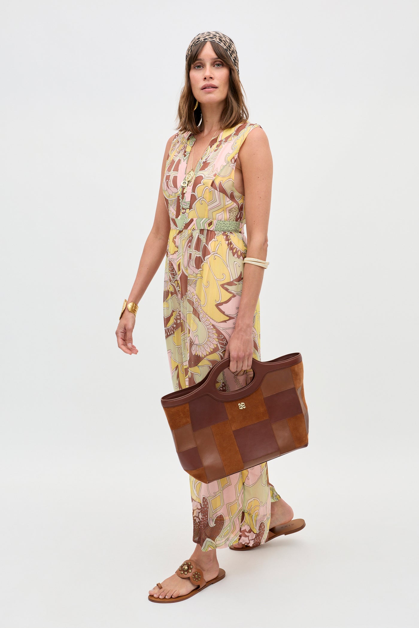 A woman stands against a plain white background, wearing the Brigid - Bianca Abstract Green sleeveless maxi dress. She holds a large brown tote bag and accessorizes with flat sandals, a headscarf, and gold jewelry.
