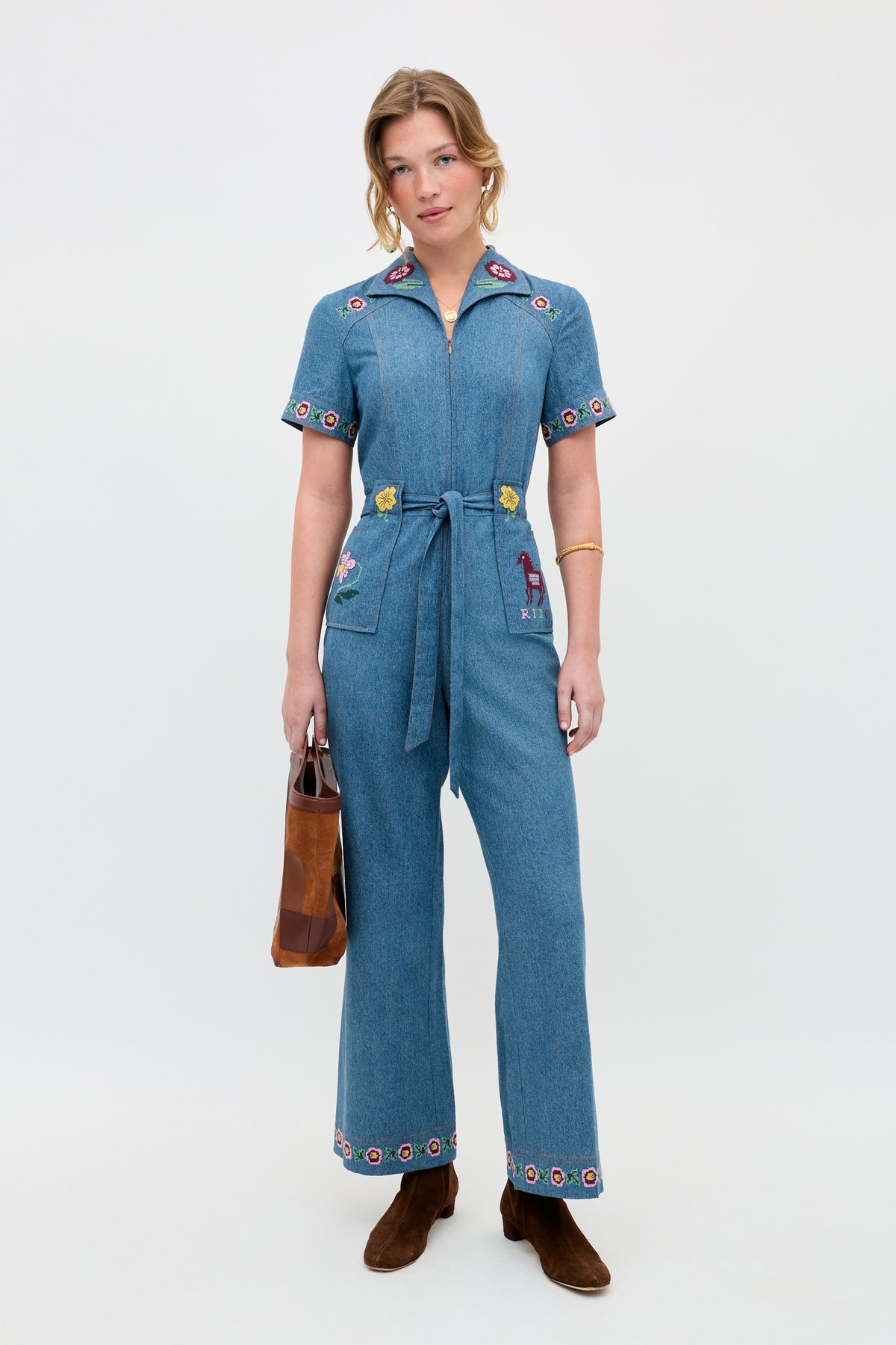 Wearing the Briana - Juliette Embroidery Blue jumpsuit, a woman with short, wavy hair and a relaxed expression stands against a plain background. She pairs the embroidered denim piece with brown boots and holds a matching brown handbag.