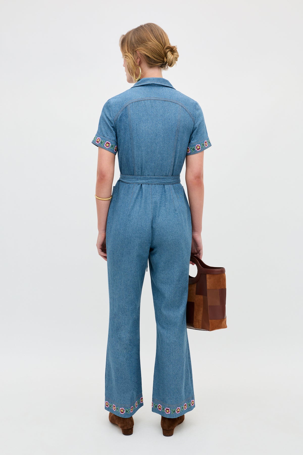 A person stands facing away, wearing the Briana - Juliette Embroidery Blue belted denim jumpsuit with colorful embroidery on the sleeves and pant legs, holding a brown handbag.