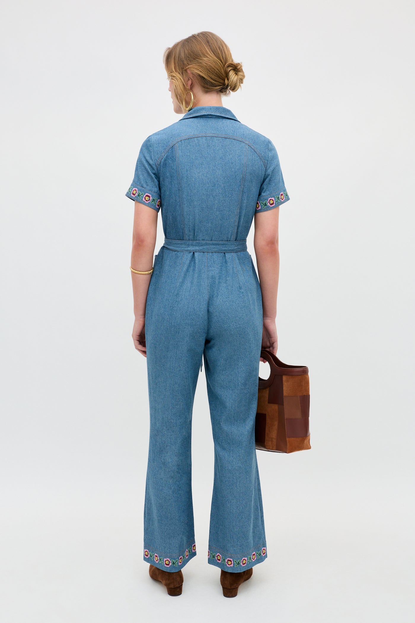 A person stands facing away, wearing the Briana - Juliette Embroidery Blue belted denim jumpsuit with colorful embroidery on the sleeves and pant legs, holding a brown handbag.