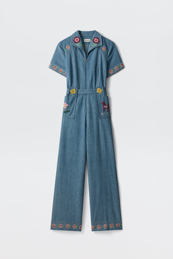 A blue denim jumpsuit, the Briana - Juliette Embroidery Blue, featuring short sleeves and wide legs with colorful floral embroidery on the collar, sleeves, pockets, and hem. One pocket is adorned with an embroidered deer. The jumpsuit is displayed against a plain white background.