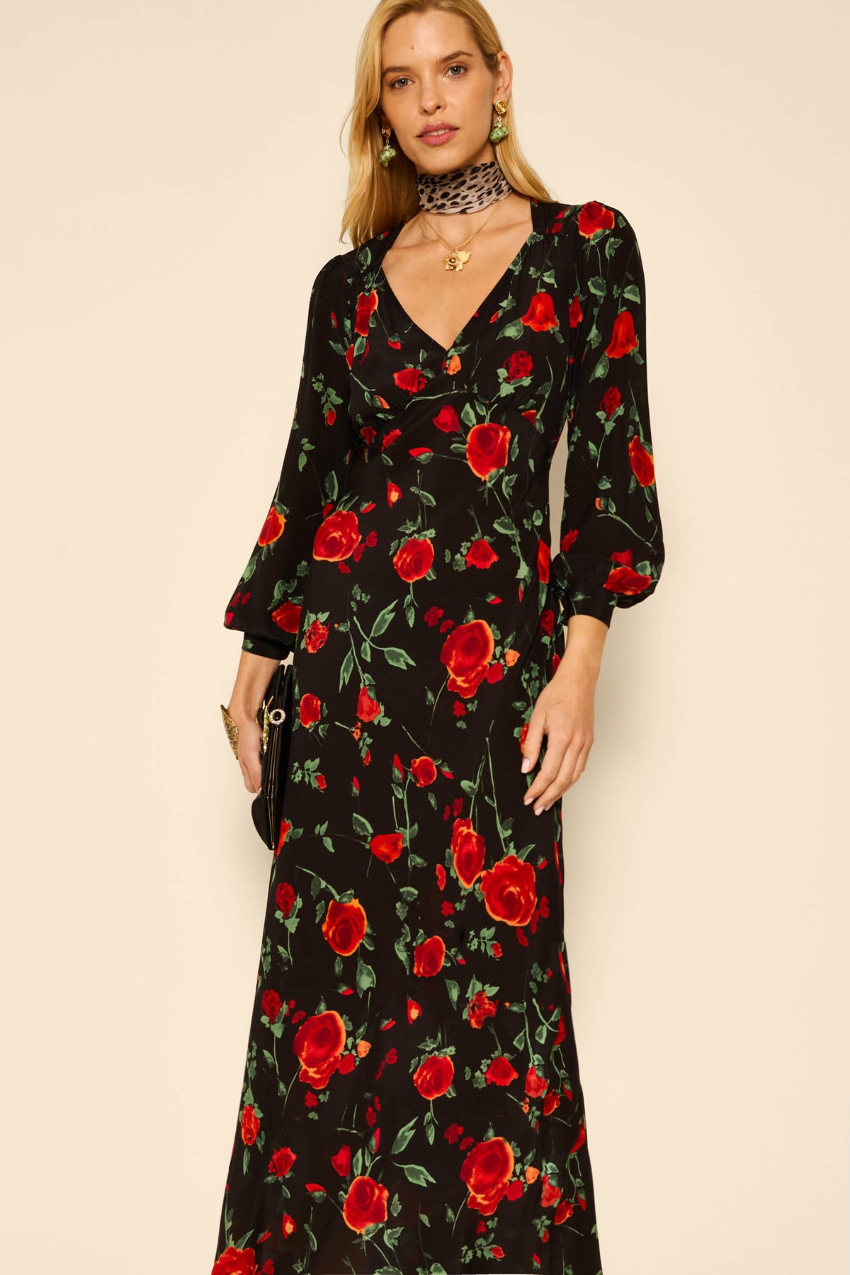 A woman stands against a plain background wearing the Mavis - Claudia Rose Black dress, featuring long sleeves and a red rose print, styled with a black purse, gold jewelry, and a black choker.