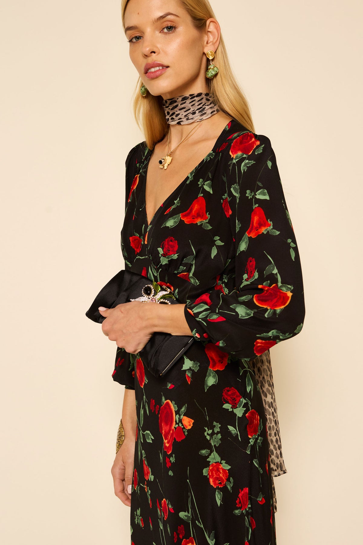 A woman stands against a plain background, wearing the Mavis - Claudia Rose Black dress with red rose print, a leopard-print scarf, gold earrings, and holding a black clutch purse.