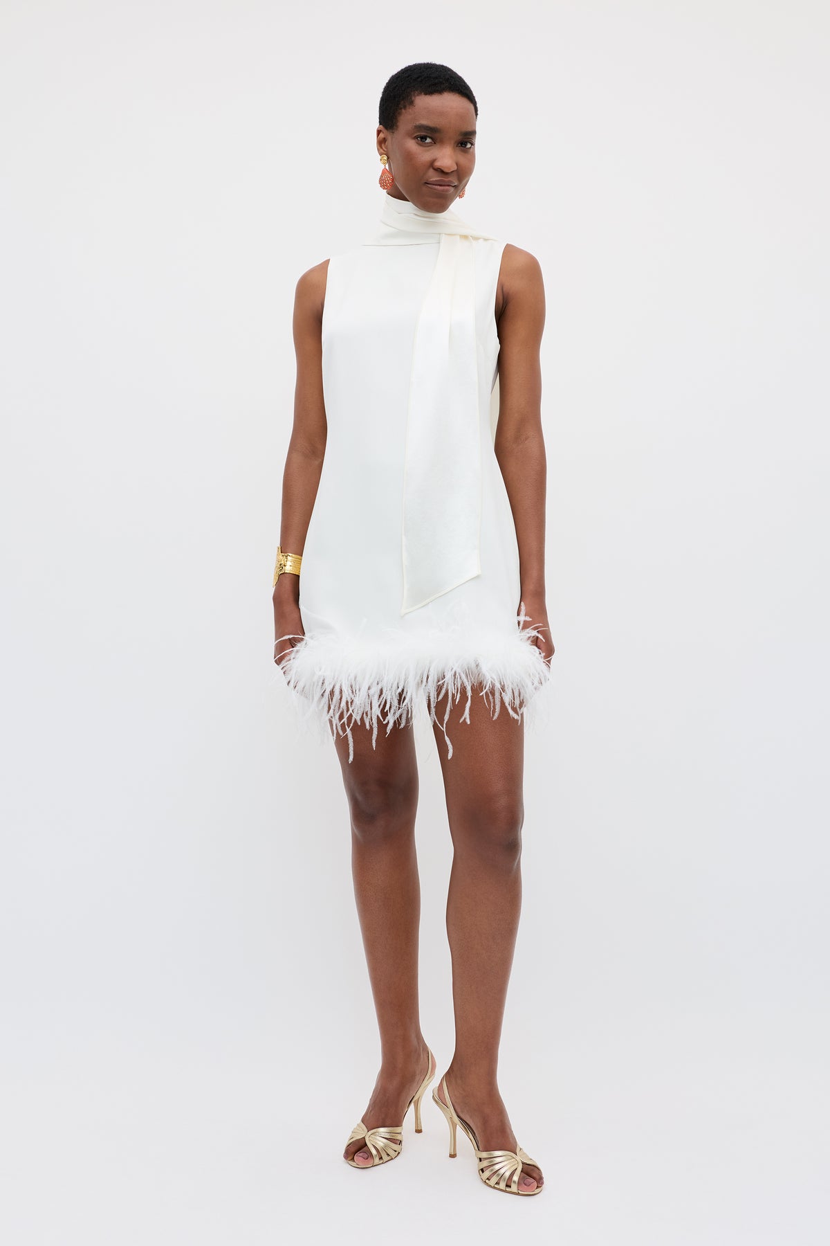 A woman wears the Hermione - Satin Back Silk Crepe sleeveless white mini dress with scarf detail and feather trim, gold heels, and jewelry against a light background—an elegant choice as a chic bridal accessory.