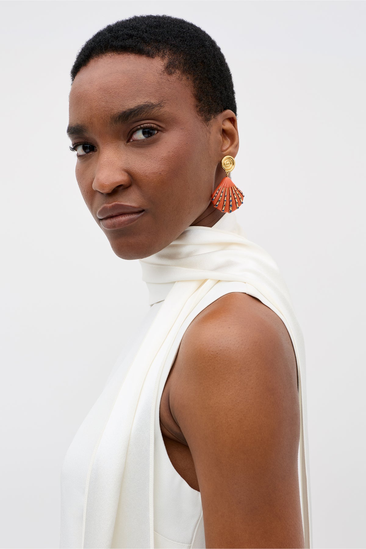 A woman with short hair wears the Hermione sleeveless white satin back silk crepe top with high neck and scarf detail, styled as a chic bridal accessory. She confidently showcases vibrant fan-shaped earrings against a plain light background.