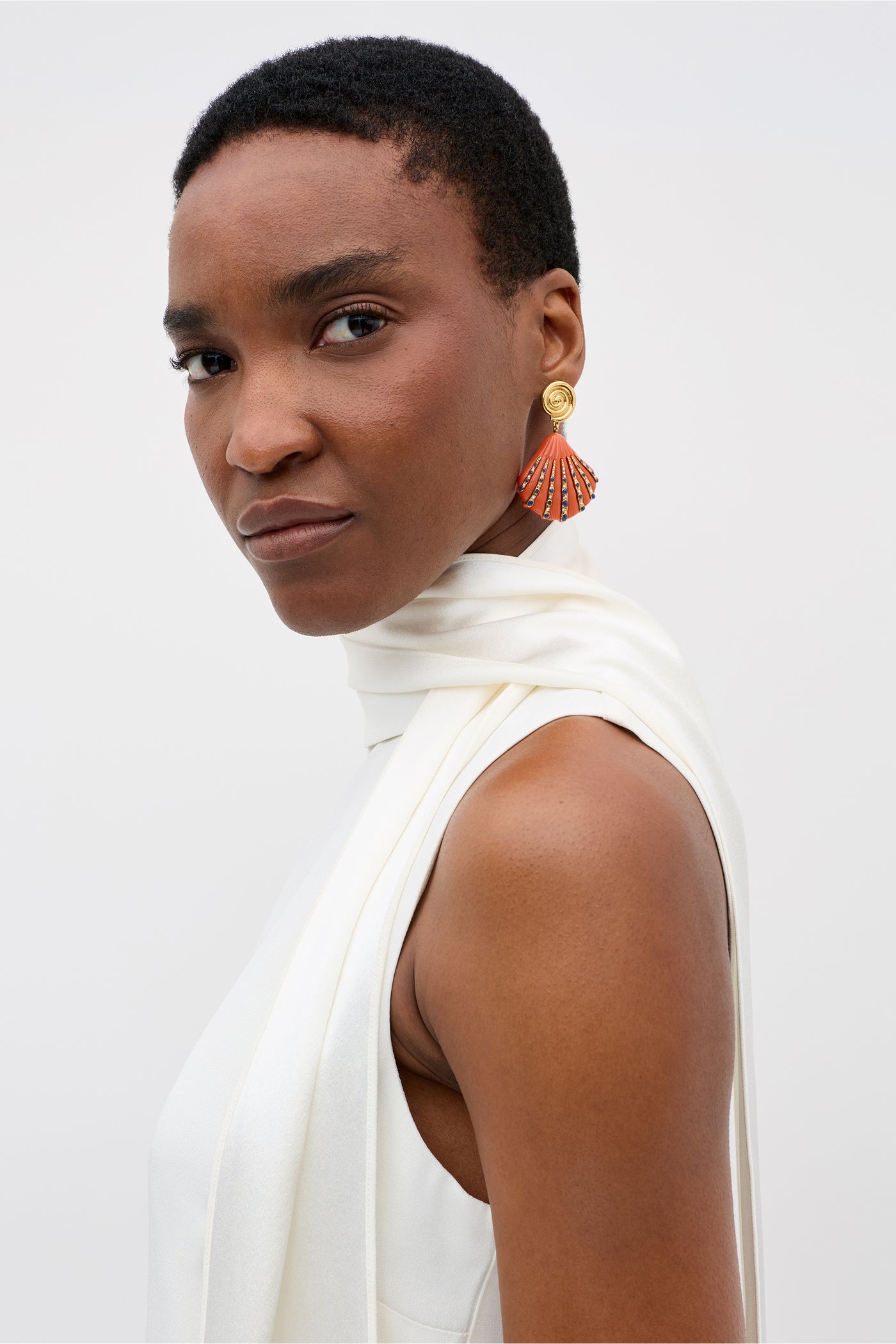 A woman with short hair wears the Hermione sleeveless white satin back silk crepe top with high neck and scarf detail, styled as a chic bridal accessory. She confidently showcases vibrant fan-shaped earrings against a plain light background.