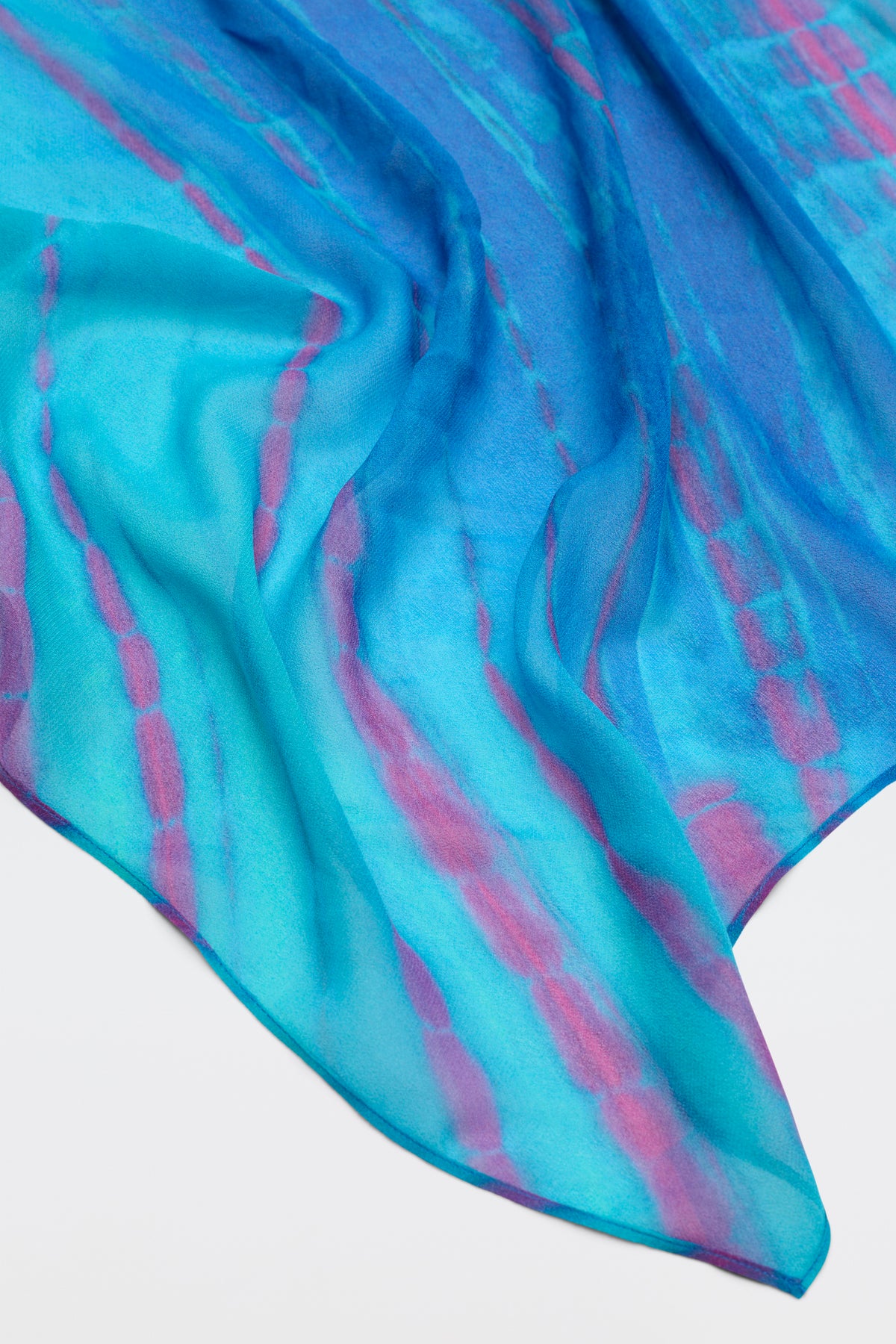 A close-up of Menno - Tie Dye Blue, featuring sheer, flowing fabric with blue, turquoise, and pink tie-dye patterns draped loosely on a light surface.
