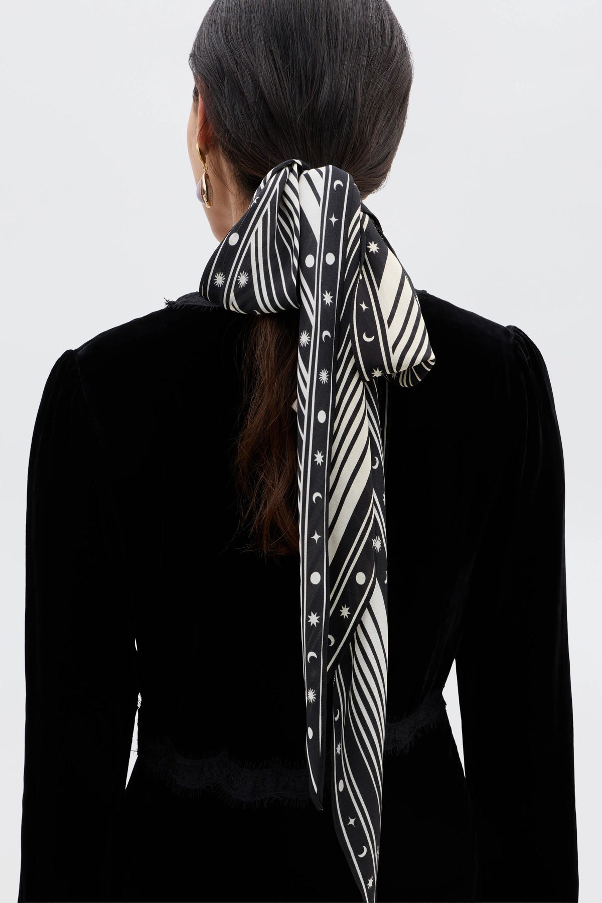 A person with long dark hair in a low ponytail, tied with the Nara - Starstruck Black scarf, pairs a black velvet top and gold hoop earrings, photographed from the back against a plain light background.