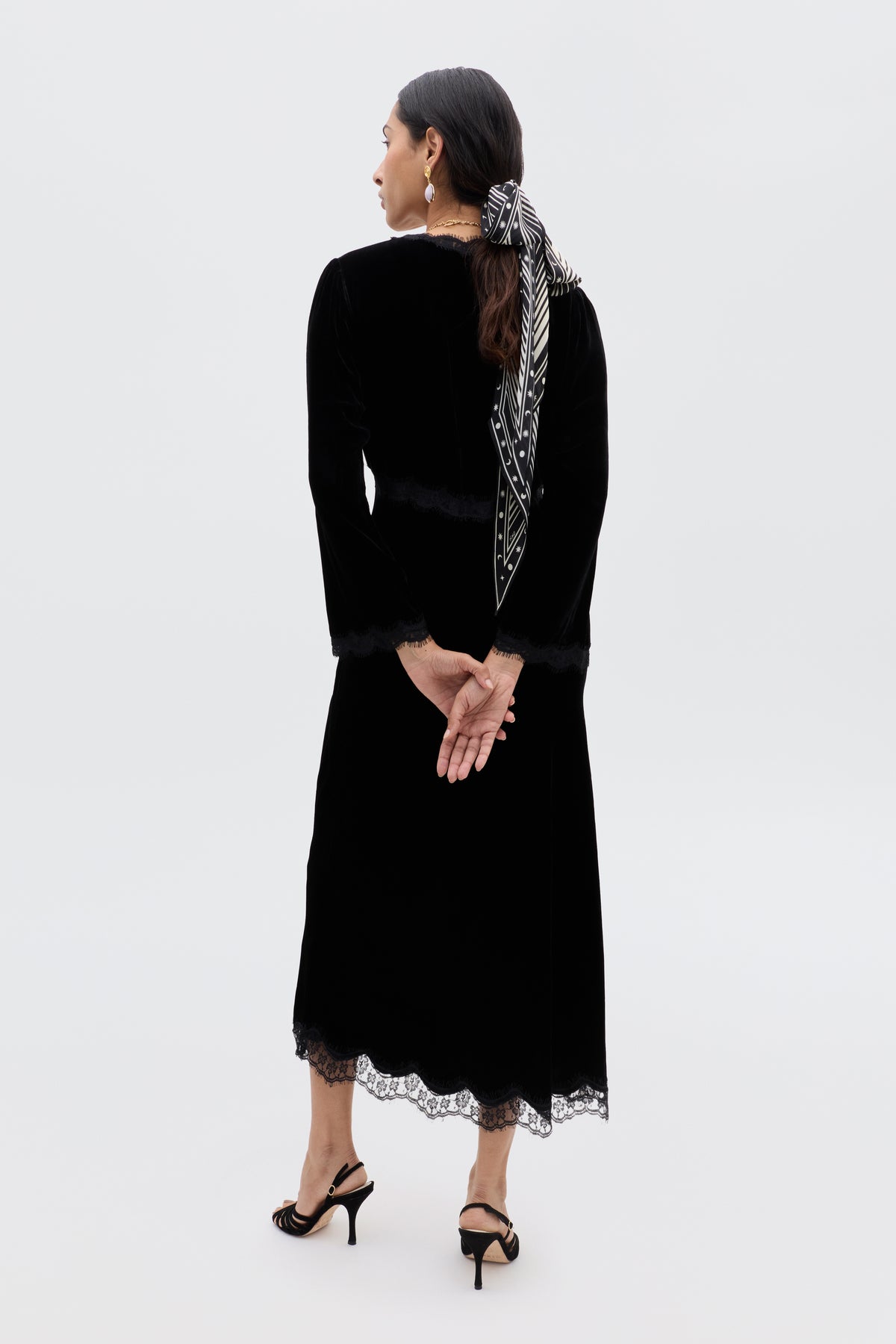 A woman faces away, hands clasped behind her back, in a long black velvet dress with lace trim, black heeled sandals, and a chic touch: the Nara - Starstruck Black scarf tied in her hair. The background is plain and light-colored.