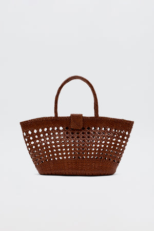 The Lucine - Tan is a brown woven straw tote bag with an openwork pattern, two handles, and a small flap closure, shown against a plain white background.