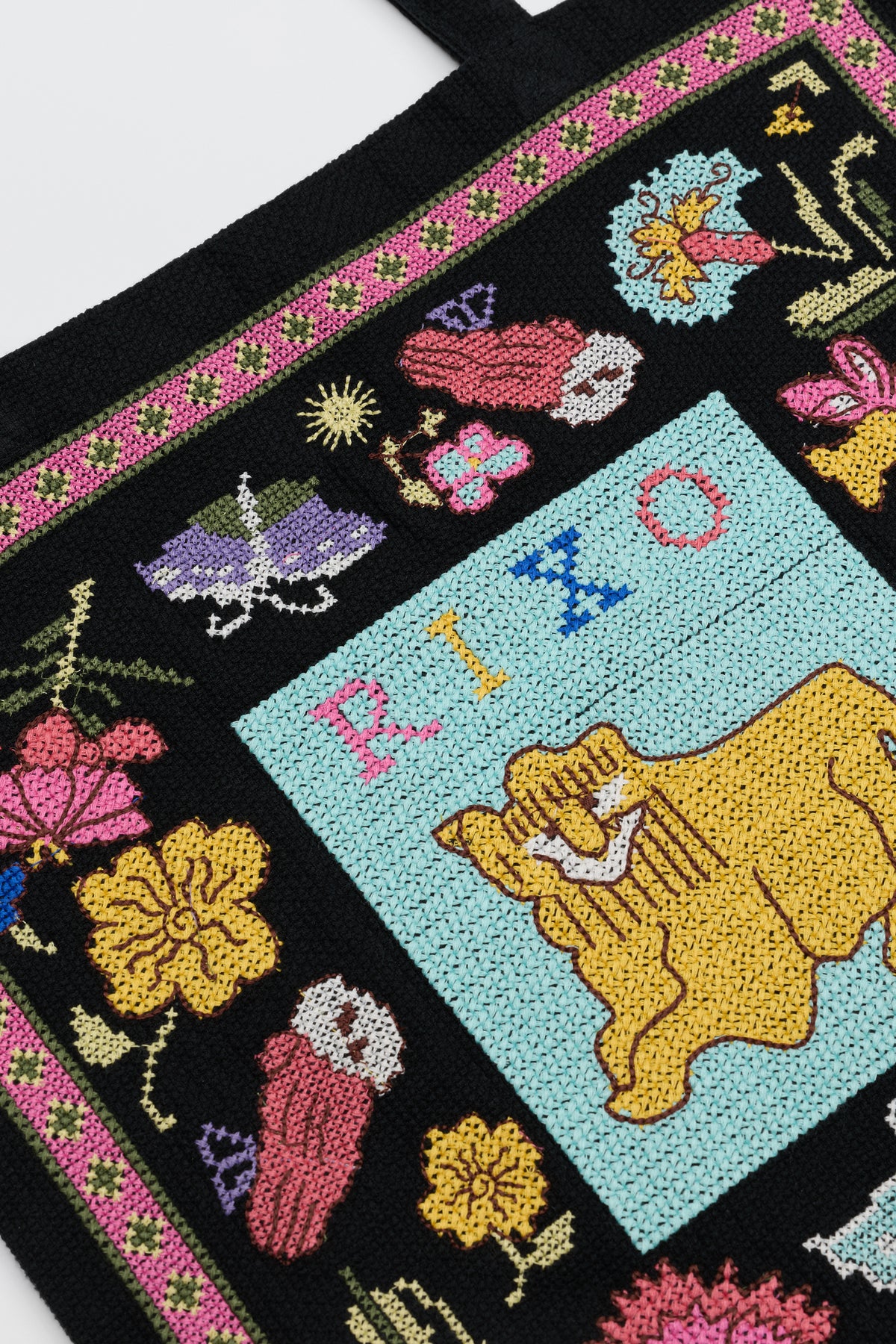 Lili – Leo Cross Stitch Embroidery Charcoal features a vibrant embroidered tapestry showcasing flowers, mushrooms, a moth, and a yellow lion set against a rich blue background. Above the lion, “RIZO” is stitched in multicolored letters, while the border is adorned with pink and green floral patterns.