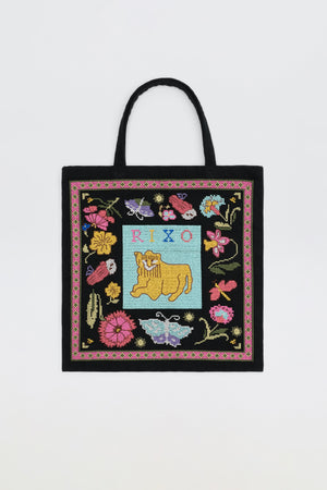 A charcoal Lili - Leo Cross Stitch Embroidery tote bag featuring colorful embroidered flowers, butterflies, and a yellow cow in the center, with “RIXO” stitched above the cow on a blue background.