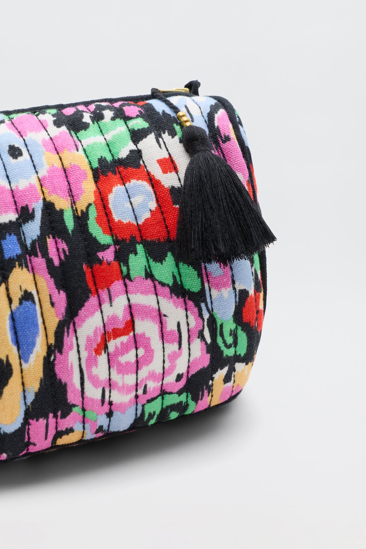 A close-up of the Jadina - Ikat Bloom Charcoal clutch, featuring pleated fabric with a colorful floral pattern and a black tassel zipper pull, set against a plain light background.