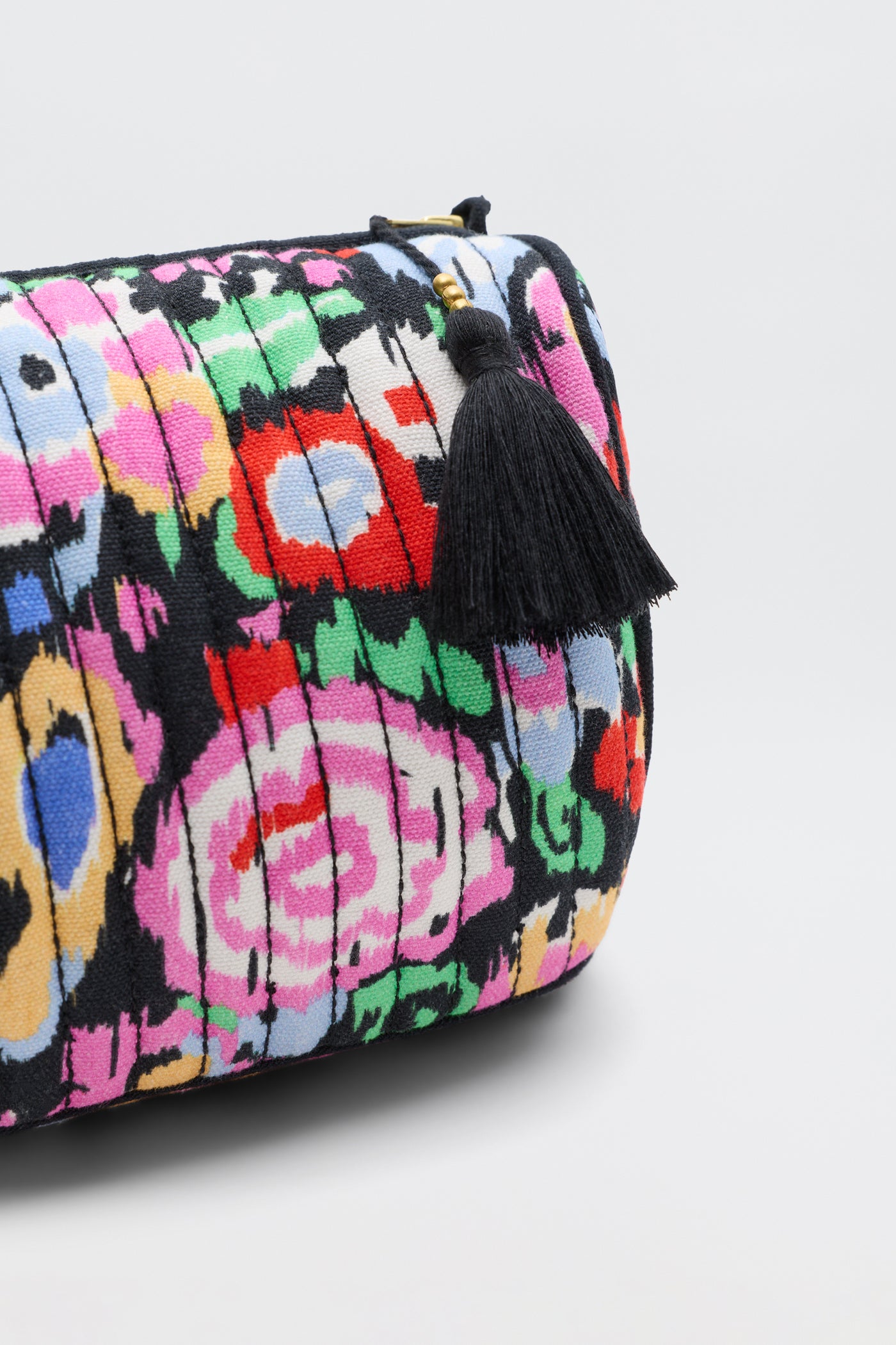 A close-up of the Jadina - Ikat Bloom Charcoal clutch, featuring pleated fabric with a colorful floral pattern and a black tassel zipper pull, set against a plain light background.
