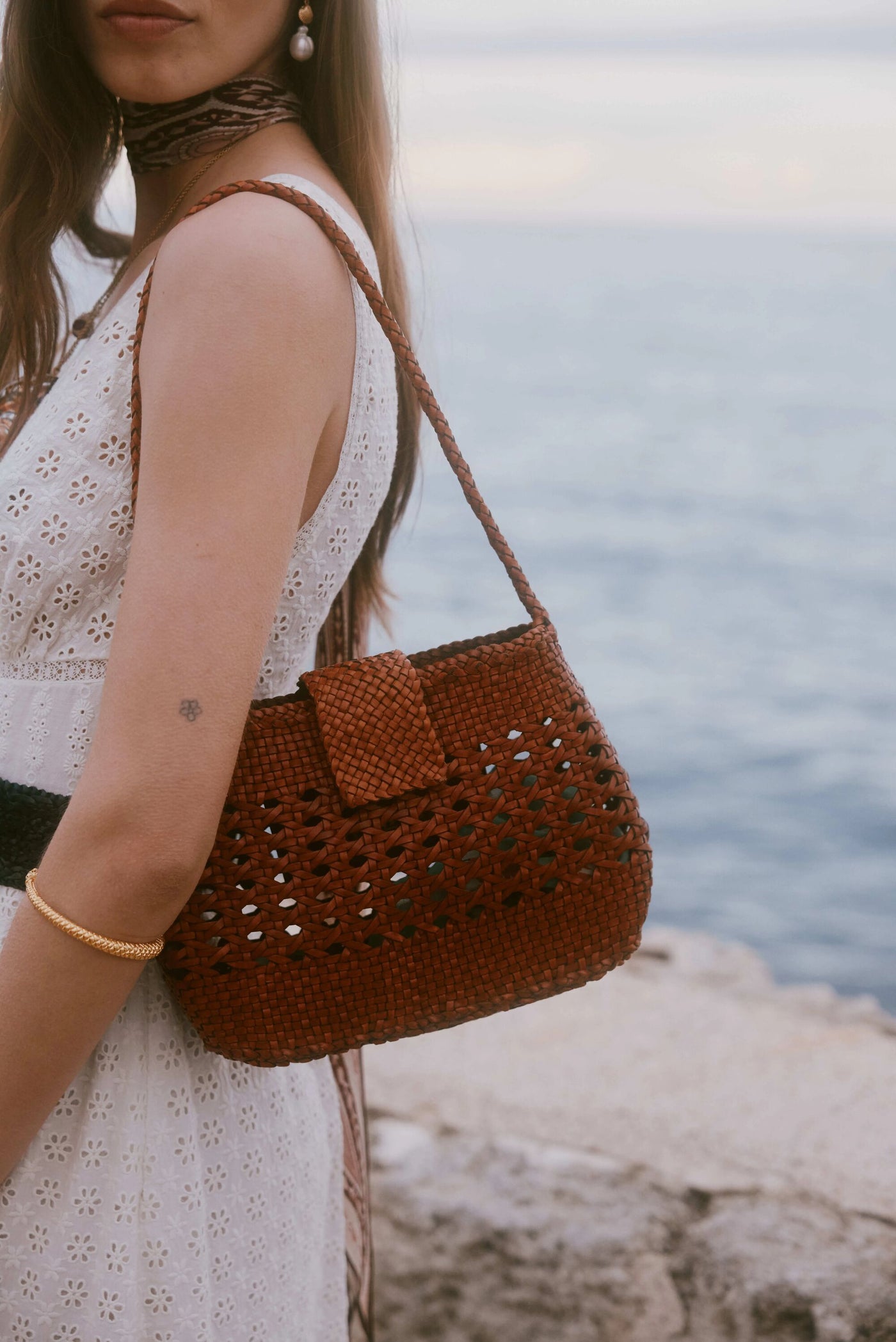 A woman in a sleeveless white eyelet dress and gold bracelet stands by the water, carrying the Amoria - Tan woven bag. Only her upper body is shown, with the sea as the backdrop.
