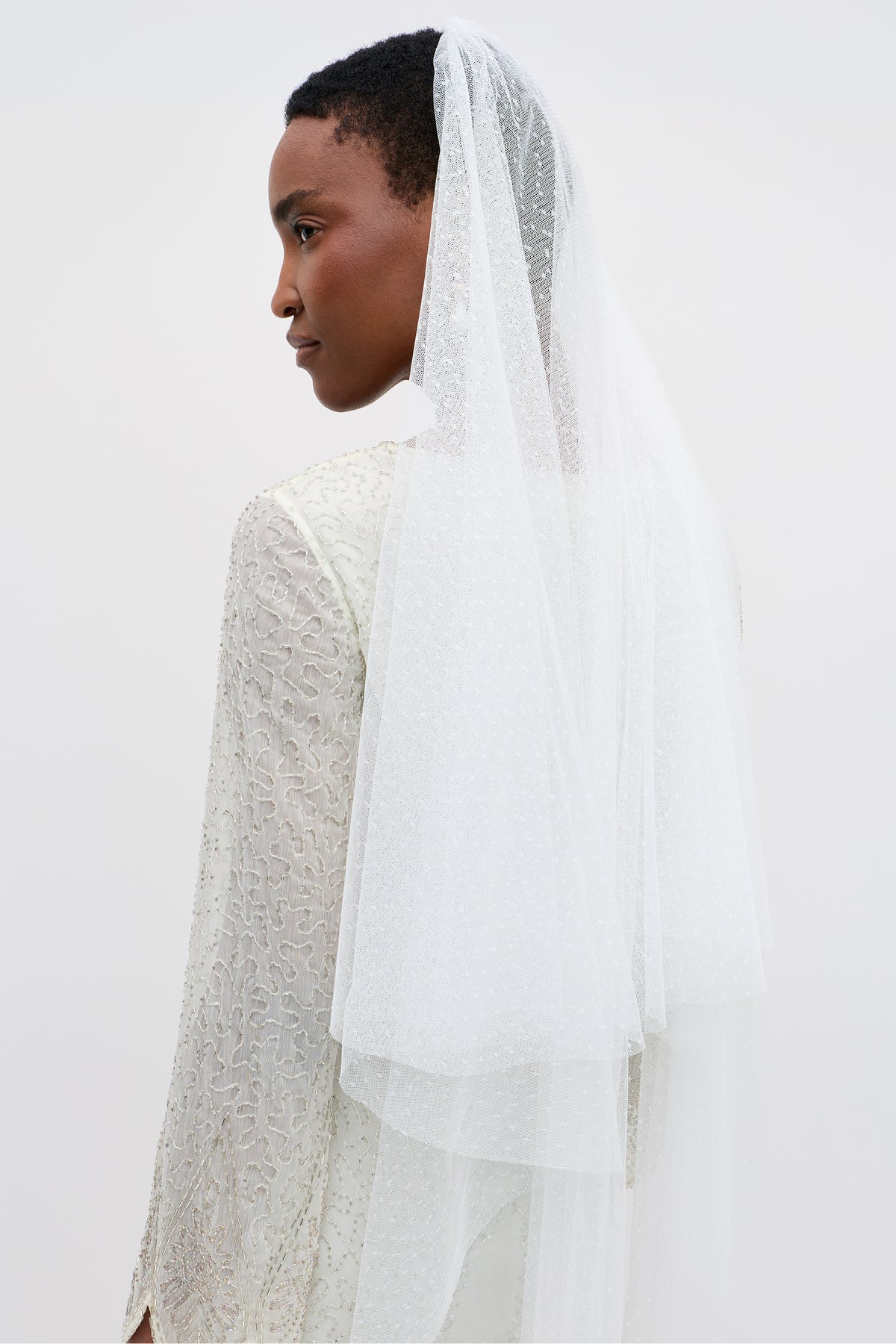 A woman in a textured white wedding dress with a dramatic train and the Lavina - Ivory Veil stands against a plain light background, shown in profile facing left.