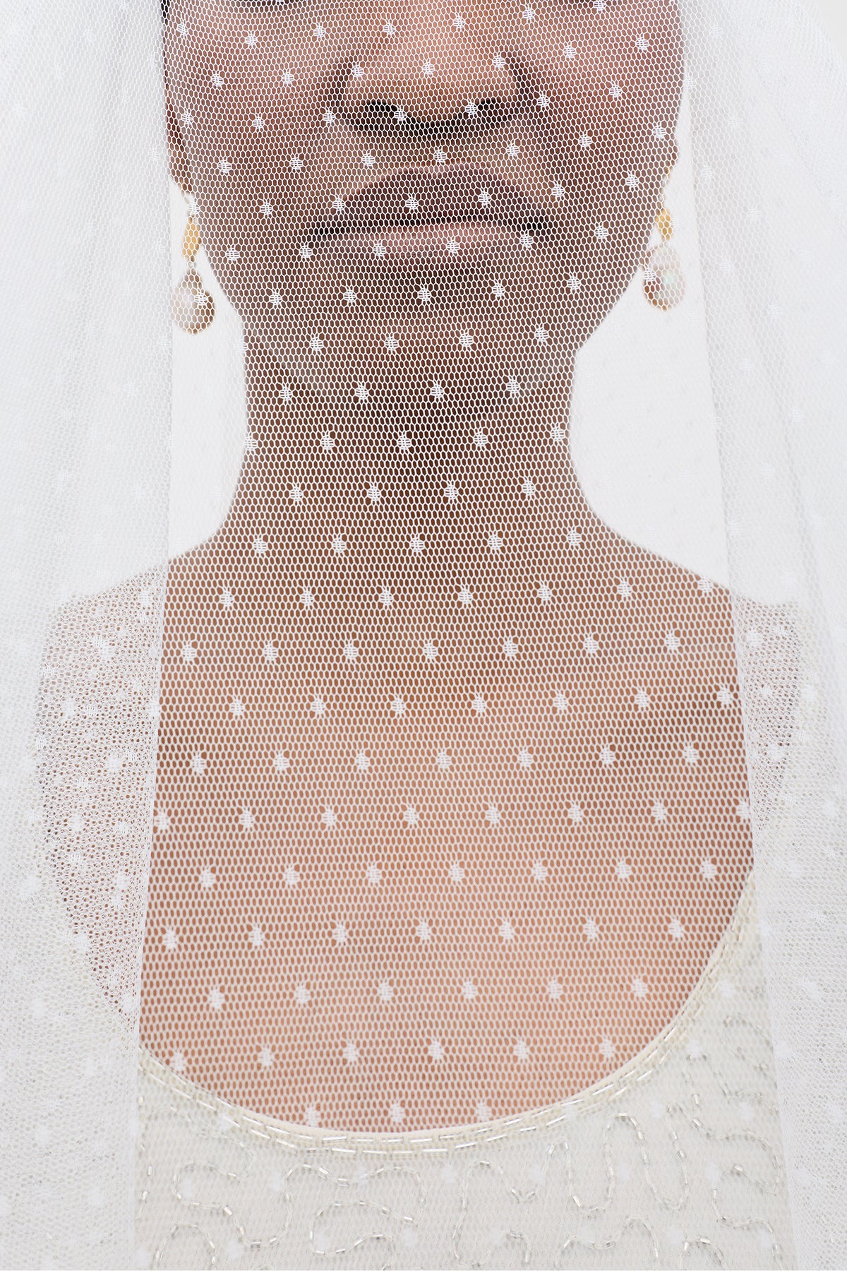 A close-up of a bride wearing the Lavina - Ivory Veil, featuring ivory polka-dotted mesh, with her lower face, neck, and upper chest visible. She also wears a white dress and pearl earrings.