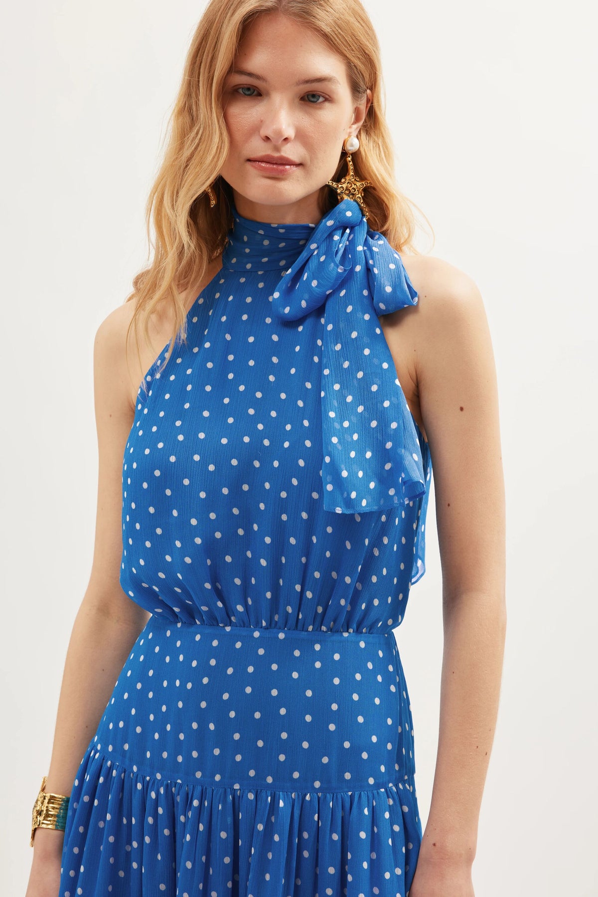 A woman models the Abi - Layla Dot Blue dress, a sleeveless blue georgette piece with white polka dots and a high neck bow, styled with gold jewelry—ideal for occasionwear or as an elegant bridesmaid’s dress.