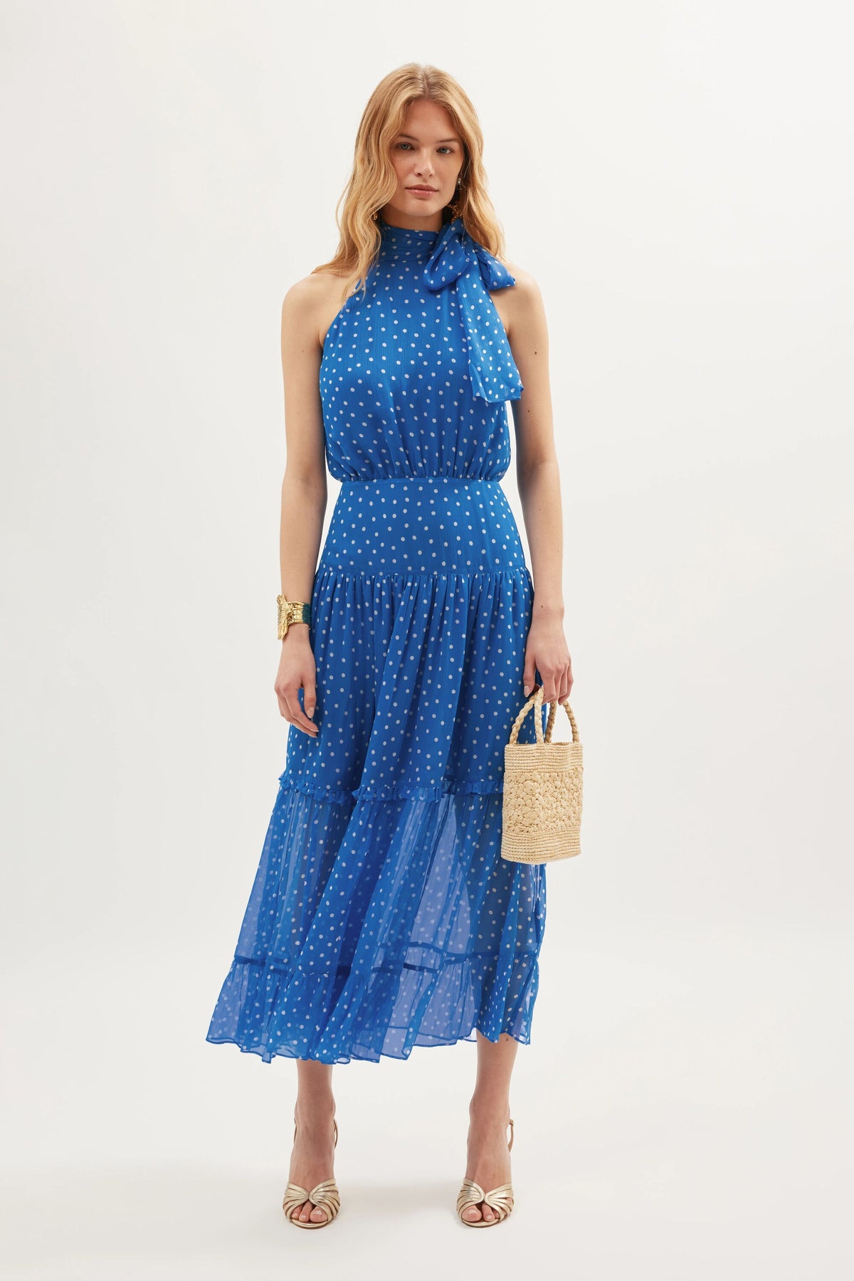 A woman stands against a white background wearing the sleeveless Abi - Layla Dot Blue dress with white polka dots, styled with strappy heels, a gold bracelet, and a woven handbag—ideal for occasionwear or as a bridesmaids dress.