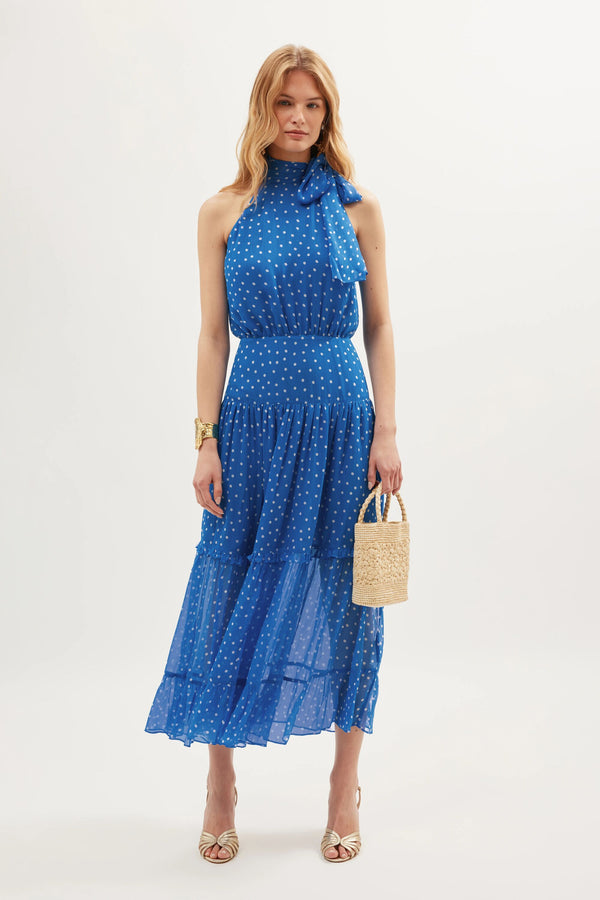 A woman stands against a white background wearing the sleeveless Abi - Layla Dot Blue dress with white polka dots, styled with strappy heels, a gold bracelet, and a woven handbag—ideal for occasionwear or as a bridesmaids dress.
