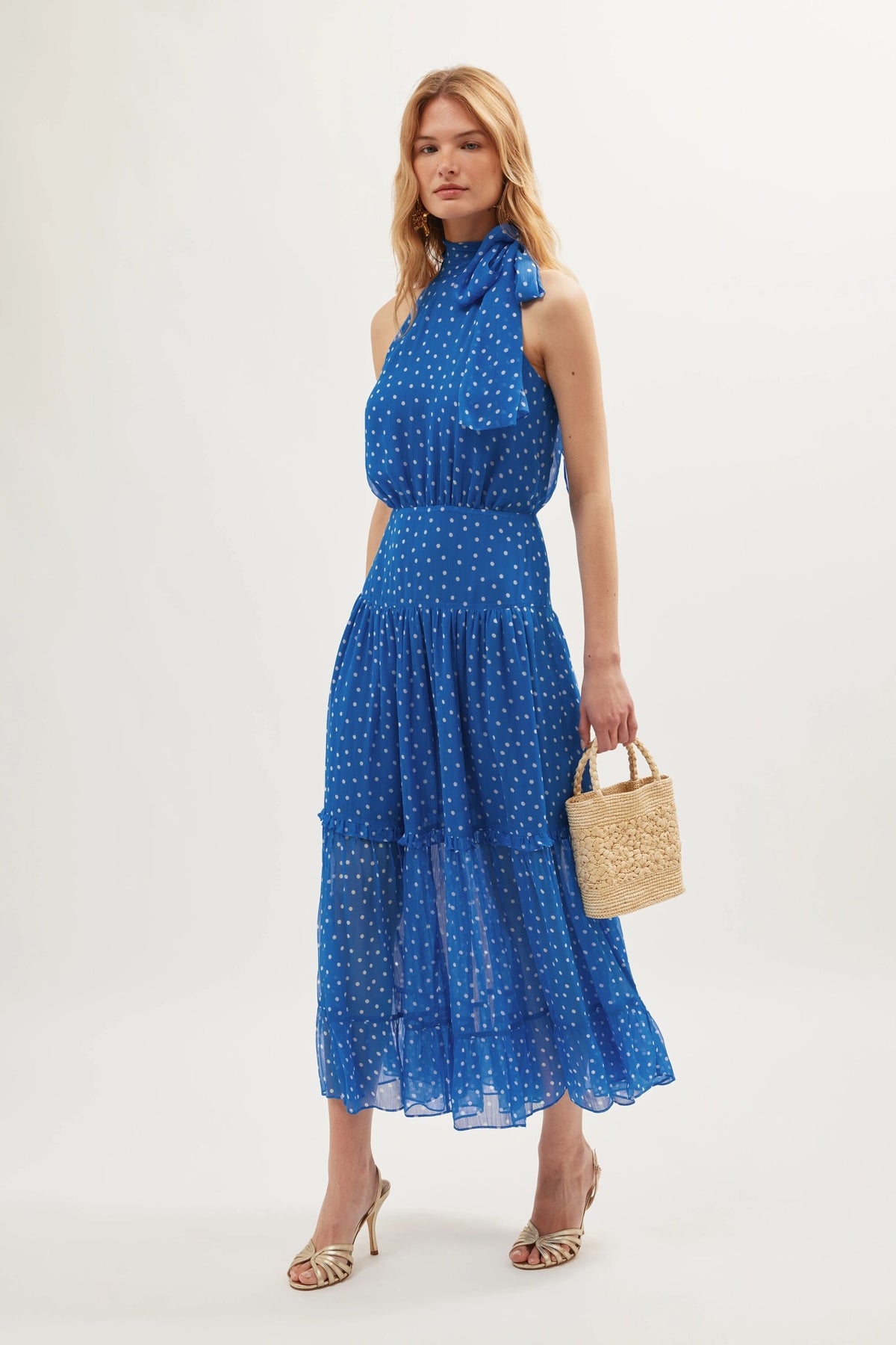 A woman in the Abi - Layla Dot Blue sleeveless georgette dress, featuring white polka dots and a neck bow, carries a small straw handbag and wears strappy heeled sandals—an ideal look for occasionwear.