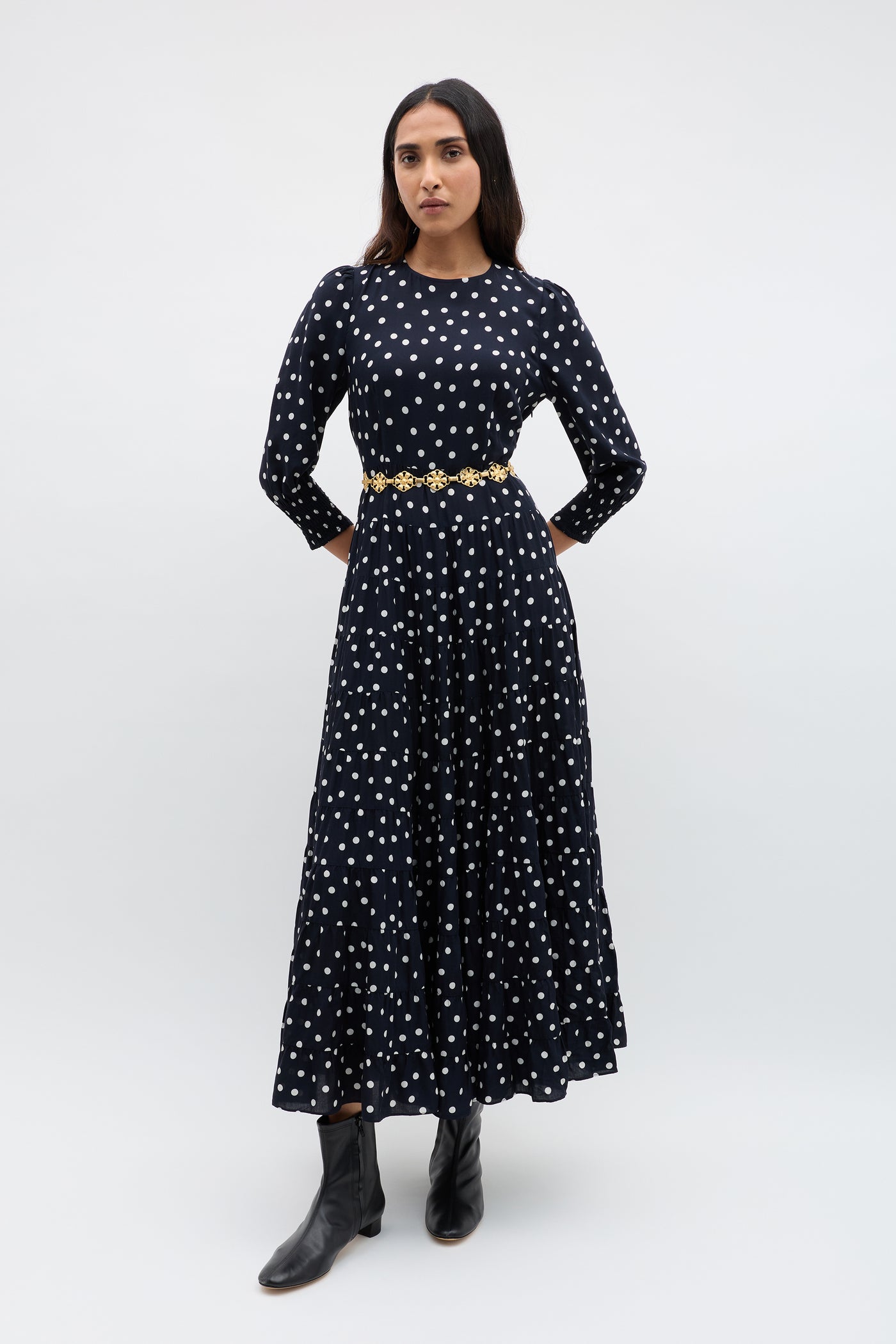 A woman in a long-sleeved, black RIXO polka-dot maxi dress, the Tiana - Gold daisy-chain belt, and black ankle boots stands against a plain, light background.