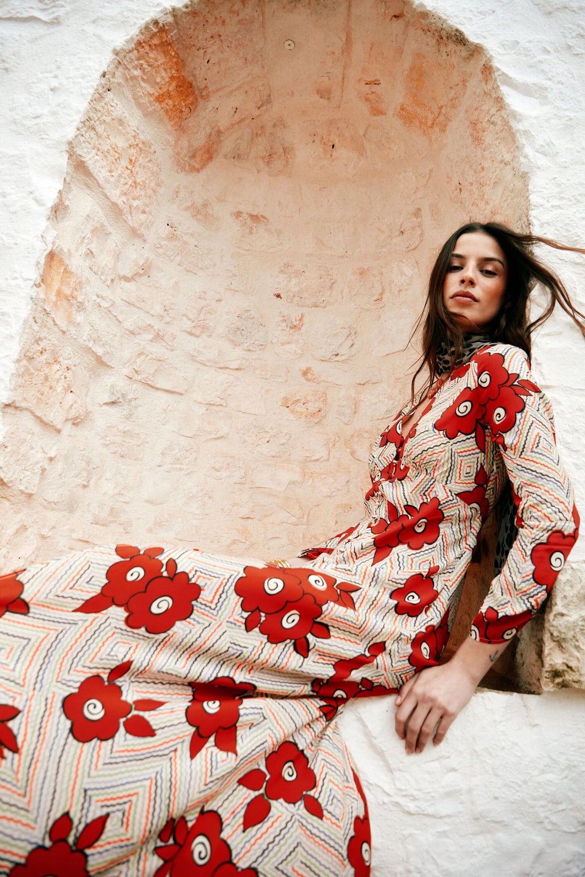 A woman in the Katie - Milos Floral Cream dress with a bold red floral print leans against a rustic, pale stone wall, her windblown hair framing her face as she looks at the camera.