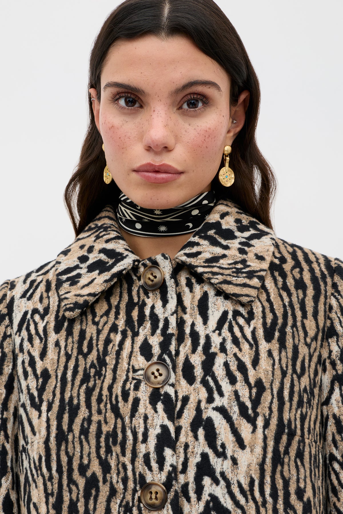 A woman with long dark hair and freckles wears the Milly - Bohemia Leopard mid-length coat with large buttons, gold drop earrings, and a black-and-white scarf around her neck, standing against a plain light background.