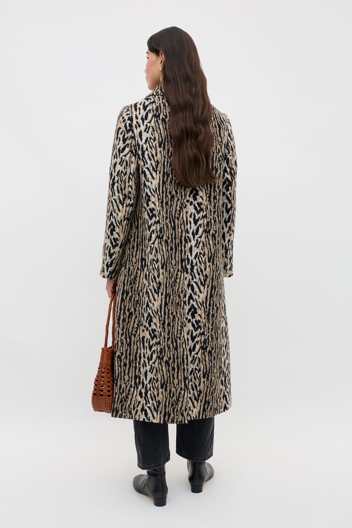 A person with long brown hair stands facing away, wearing the Milly - Bohemia Leopard mid-length coat, black pants and boots, holding a brown woven bag against a plain light background.